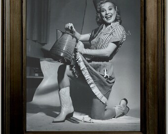 May include: A woman wearing a cowboy hat and a striped shirt is kneeling and holding a metal pitcher. She is smiling and looking at the camera. The background is a light gray wall.