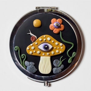 May include: A round, silver compact mirror with a black background. The mirror features a whimsical design of a yellow mushroom with an eye, a snail, a flower, and a yellow sun. The design is made of clay.