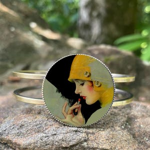May include: A gold-tone metal bracelet with a circular charm featuring a vintage illustration of a woman applying lipstick. The woman is wearing a yellow hat and has red lipstick on. The bracelet is resting on a rock.