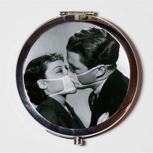 May include: A black and white photo of a couple kissing while wearing face masks. The photo is printed on a round, silver compact mirror.