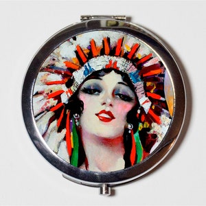 May include: A silver compact mirror with a colorful image of a woman wearing a red, white, and green headdress. The woman has dark hair and is wearing makeup.