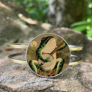 May include: A gold-toned metal bracelet with a circular charm featuring a green frog playing a banjo. The frog has green spots and is set against a cream-colored background. The bracelet is resting on a textured rock surface.