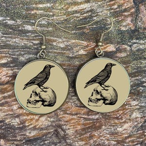 May include: A pair of circular earrings. Each earring displays a black raven perched on a skull, set against a tan backdrop. The raven and skull are in black and white. The hooks are gold-coloured.