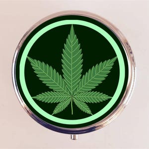 May include: A round silver pill box with a green marijuana leaf design inside a light green circle on a black background.
