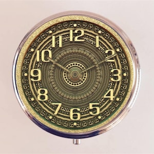 May include: A gold-colored clock face with a circular design of concentric circles and a black clock hand. The clock face has Roman numerals for the hours and Arabic numerals for the minutes.