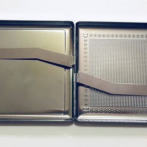 Puede incluir: Una cajetilla de cigarrillos de metal plateado con una tapa con bisagras y un patr&oacute;n texturizado en el exterior. La cajetilla est&aacute; abierta y el interior est&aacute; vac&iacute;o.