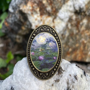 May include: An oval-shaped, bronze-colored metal pin with a detailed design. The pin features a painting of water lilies in a pond, with a blue and purple sky in the background.