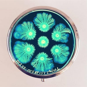 May include: A round, silver compact mirror with a black background and a design of six green and blue crystal formations. The text "7.95 ml" and "26.67.CO.23.2" is printed on the bottom of the mirror.