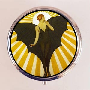 May include: A round, silver-colored trinket box with a vintage illustration of a woman with bat wings against a yellow sunburst background.