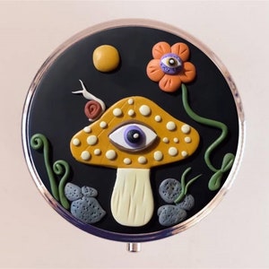 May include: A round, black compact mirror with a whimsical design featuring a yellow mushroom with a purple eye, a red flower with a purple eye, and green vines. The background is black.