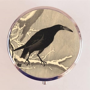 May include: A round, silver compact mirror featuring a black crow perched on a branch. The crow is depicted in a detailed, artistic style against a muted background of beige and gray tones. The compact has a shiny, metallic border.