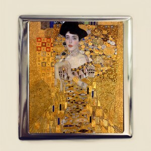 May include: A silver metal case with a portrait of a woman in a gold and black patterned dress. The woman has dark hair and is wearing a pearl necklace. The background is a gold and black geometric pattern.