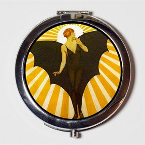 May include: A compact mirror with a vintage illustration of a woman with wings against a yellow and white sunburst background.