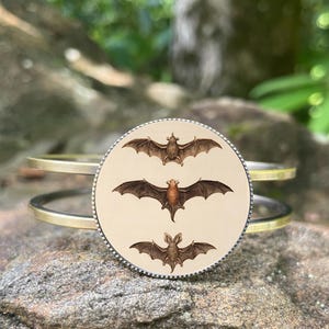 May include: A gold and silver-toned cuff bracelet featuring a circular pendant with three brown bat illustrations. The bats are depicted with outstretched wings against a cream-colored background. The bracelet is resting on a rock.