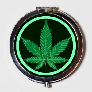 May include: A silver compact mirror with a black background and a green marijuana leaf design inside a light green circle.