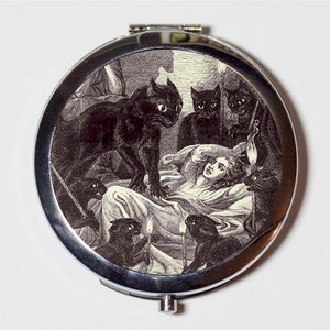 May include: A silver compact mirror with a black and white illustration of a woman surrounded by black cats. The illustration is in a vintage style.