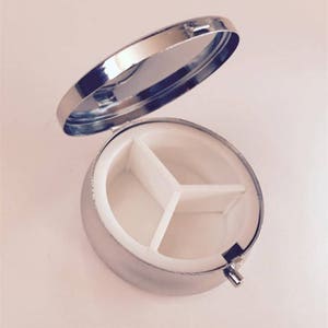 May include: A small, round, silver pill box with a hinged lid. The interior is divided into three white compartments. The lid is open, revealing the inside. The pill box is designed for organizing and carrying medication or vitamins.