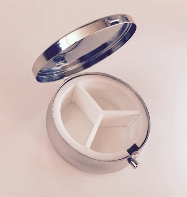 May include: A small, round, silver pill box with a hinged lid. The interior is divided into three white compartments. The lid is open, revealing the inside. The pill box is designed for organizing and carrying medication or vitamins.