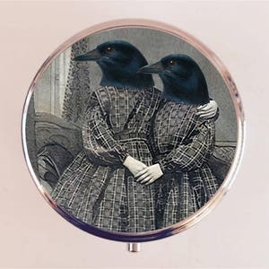 May include: A vintage-style compact mirror featuring a black and white image of two crows with human bodies wearing plaid dresses. The crows are looking at the camera and have their arms around each other.