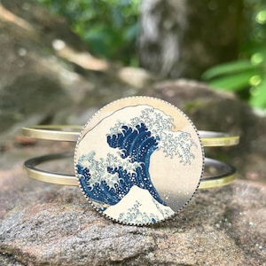 May include: A gold-toned metal cuff bracelet featuring a circular pendant with a print of Hokusai's "The Great Wave off Kanagawa." The artwork depicts a large blue wave with white foam, a mountain, and a light background.