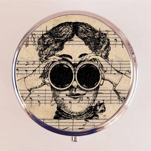 May include: A vintage-style compact mirror with a black and white illustration of a person looking through binoculars. The background is a sheet of music.