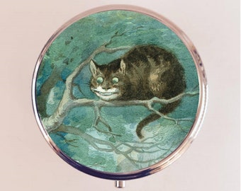 May include: A round, silver compact mirror with a vintage illustration of a Cheshire Cat with a wide grin, sitting on a branch in a forest setting. The cat has green eyes and is depicted in a whimsical style.