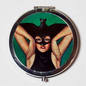 May include: A silver compact mirror with a round, green background featuring a woman wearing a black bat costume. The woman has her arms raised above her head, and the bat wings are spread out behind her.