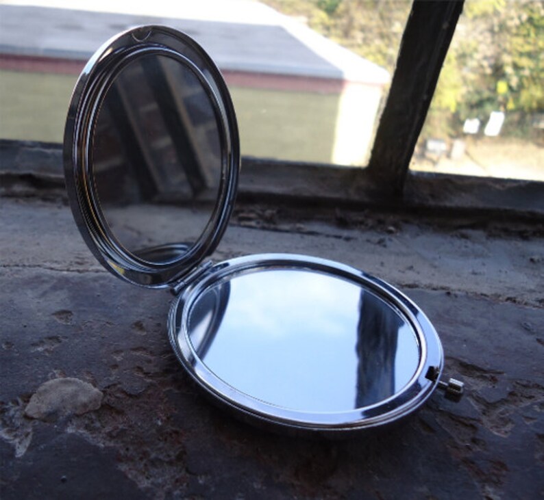 May include: Open, silver compact mirror with two reflective surfaces. The compact is oval-shaped and has a hinged design. The mirror is open, revealing the reflective surfaces. The compact is on a textured surface.