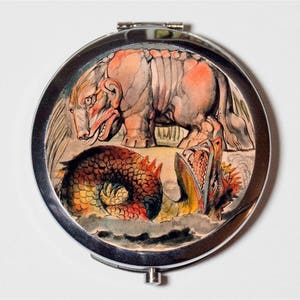 May include: A silver compact mirror with a colorful illustration of a mythical creature with a large mouth and a scaly body, and a pink, four-legged creature with a long snout.