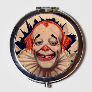 May include: A silver compact mirror with a vintage illustration of a smiling clown with red hair and a white collar. The clown has a missing tooth and is wearing black makeup around the eyes.