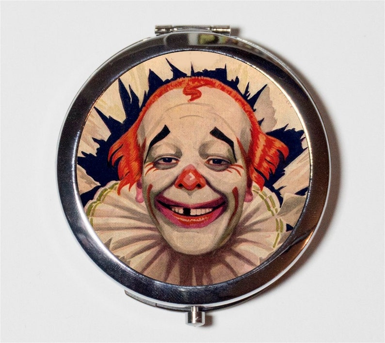 May include: A silver compact mirror with a vintage illustration of a smiling clown with red hair and a white collar. The clown has a missing tooth and is wearing black makeup around the eyes.