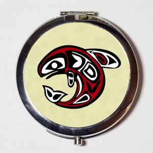 May include: A compact mirror with a silver metal frame and a round, cream-colored lid featuring a stylized red, black, and white salmon design.