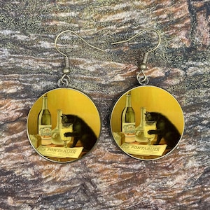 May include: A pair of round earrings featuring a vintage-style illustration. Each earring displays a black cat reaching for a glass, with bottles and the text "PONTARLIER" on a yellow background. The earrings have silver-colored hooks.