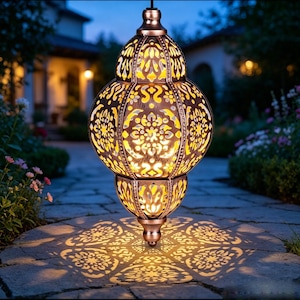 May include: A hanging metal lantern with a bronze finish and intricate cut-out patterns. The lantern emits a warm yellow light from within, casting detailed floral and geometric shadows onto the stone ground surface below it.