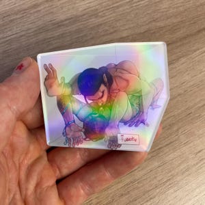 May include: A trapezoidal acrylic charm with a holographic finish featuring an illustration of a figure with dark hair and pointed ears in a crouched pose. The artwork includes a small white rectangular label with the handwritten text For Robert.