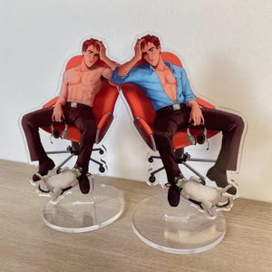 May include: Two acrylic standees featuring a character with short red hair sitting in an orange office chair. One version shows the character shirtless, the other wears a blue button-down shirt. Both hold headphones and have a small dog at their feet.