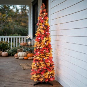May include: A tall, slender, conical artificial tree decorated with vibrant red, orange, and yellow maple leaves. The tree is illuminated by small, warm white string lights woven throughout the foliage and stands on a black metal base.