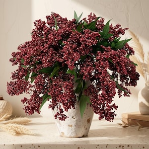 May include: A dense arrangement of deep red berry clusters with green leaves displayed in a textured, off-white ceramic vase. The botanical stems feature numerous small, round berries in a rich burgundy hue, creating a full, rounded shape.