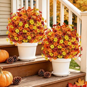 May include: Two white plastic pots each containing a dense, spherical arrangement of artificial flowers in shades of yellow, orange, and deep red. The pots are placed on wooden steps alongside several brown pinecones and orange pumpkins.