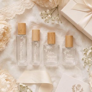 以下が含まれることがあります： 竹製の蓋が付いた4本の透明なガラス瓶。白い花柄と「Mrs. Seraphina」の文字がプリントされています。背の高い長方形のボトル2本と、背の低い正方形のボトル2本が、クリーム色の花が添えられた白い布の上に配置されています。