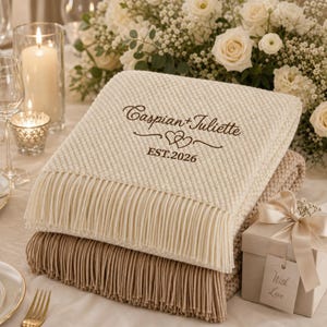 May include: A cream-coloured textured throw blanket with fringe, embroidered with the brown text Caspian + Juliette, a heart icon, and EST. 2026. The blanket is folded atop a tan blanket with fringe, next to a gift box with a ribbon and tag.