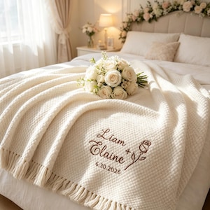 Könnte beinhalten: Eine cremefarbene Strickdecke mit Fransen, die eine braune Stickerei mit dem Text Liam + Elaine sowie dem Datum 30.06.2026 und einer Strichzeichnung einer Rose aufweist. Ein Strauß weißer Rosen liegt auf der Decke.