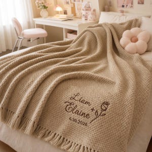 Può includere: Una coperta beige in maglia a nido d'ape con frange, caratterizzata da un ricamo marrone scuro con la scritta Liam + Elaine, un'illustrazione di una rosa e la data 30.06.2026. La coperta è adagiata su un letto con un cuscino a fiore.