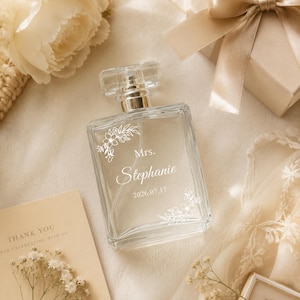 Peut inclure: Un flacon de parfum en verre transparent de forme rectangulaire avec un bouchon argenté. L'avant du flacon présente une inscription blanche gravée Mrs. Stephanie et la date 2026.07.17, ornée de délicats motifs floraux en lignes blanches.