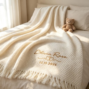 May include: A cream-coloured textured knit throw blanket with fringe edges, featuring gold embroidered text that reads Blivia Rose, a double heart icon, and the date 15.12.2026. A small brown plush teddy bear sits on the blanket.