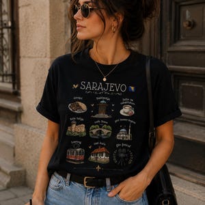 Peut inclure: Un t-shirt noir à manches courtes avec une impression graphique de monuments et d'icônes culturelles de Sarajevo, incluant les textes "SARAJEVO," "ćevapi," "Baščaršija," "latte sa rahatlukom," "žuta tabija," "vrelo Bosne," "gazi husrev-begova," "tramvaj," "inat kuća," et "film festival."