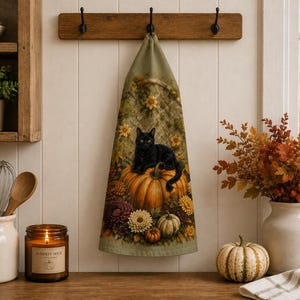 May include: A kitchen towel featuring a black cat resting on a large orange pumpkin, surrounded by autumn flowers and smaller pumpkins. The towel has a muted green background with a subtle lattice pattern and is hanging from a wooden wall hook.