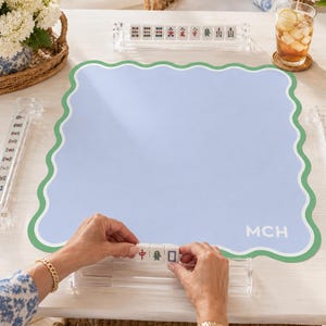 Op de afbeelding: Een vierkante lichtblauwe mahjongmat met een geschulpte groen-witte rand, voorzien van de letters MCH in de rechterbenedenhoek. De mat ligt op een tafel met vier doorzichtige acryl tegelhouders, mahjongstenen en bloemstukken.