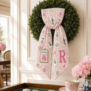 May include: A light pink fabric sash with a repeating pattern of Mahjong tiles, floral illustrations, and peacock motifs, featuring a prominent embroidered letter R and a rectangular patch with a pink monkey and the text JOKER, draped over a green wreath.
