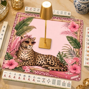 May include: A square mahjong mat featuring a pink background with a leopard wearing a pink headscarf, sunglasses, and a pearl necklace. The mat is decorated with tropical palm leaves, hibiscus flowers, and a cocktail glass. A gold lamp sits in the center.